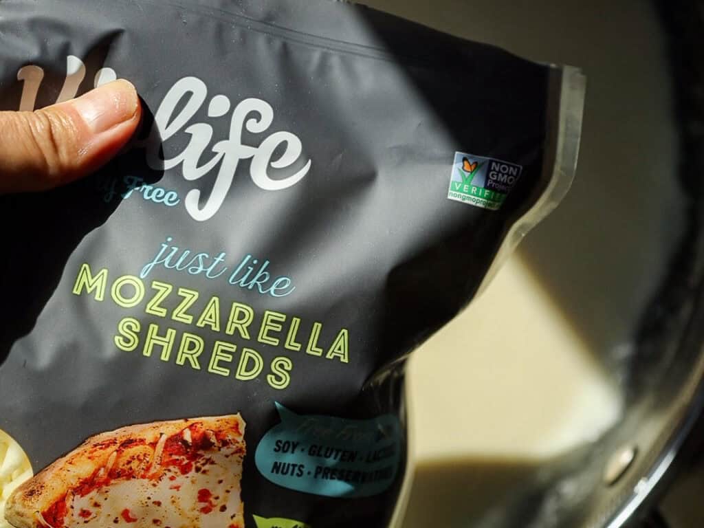 A close-up of a hand holding a black package labeled just like mozzarella shreds from a dairy-free brand. The packaging highlights non-GMO verification and is soy, gluten, lactose, nut, and preservative-free. A pizza image is visible.